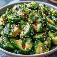 A vibrant bowl of smashed cucumber and avocado salad, drizzled with sesame dressing and sprinkled with toasted seeds.